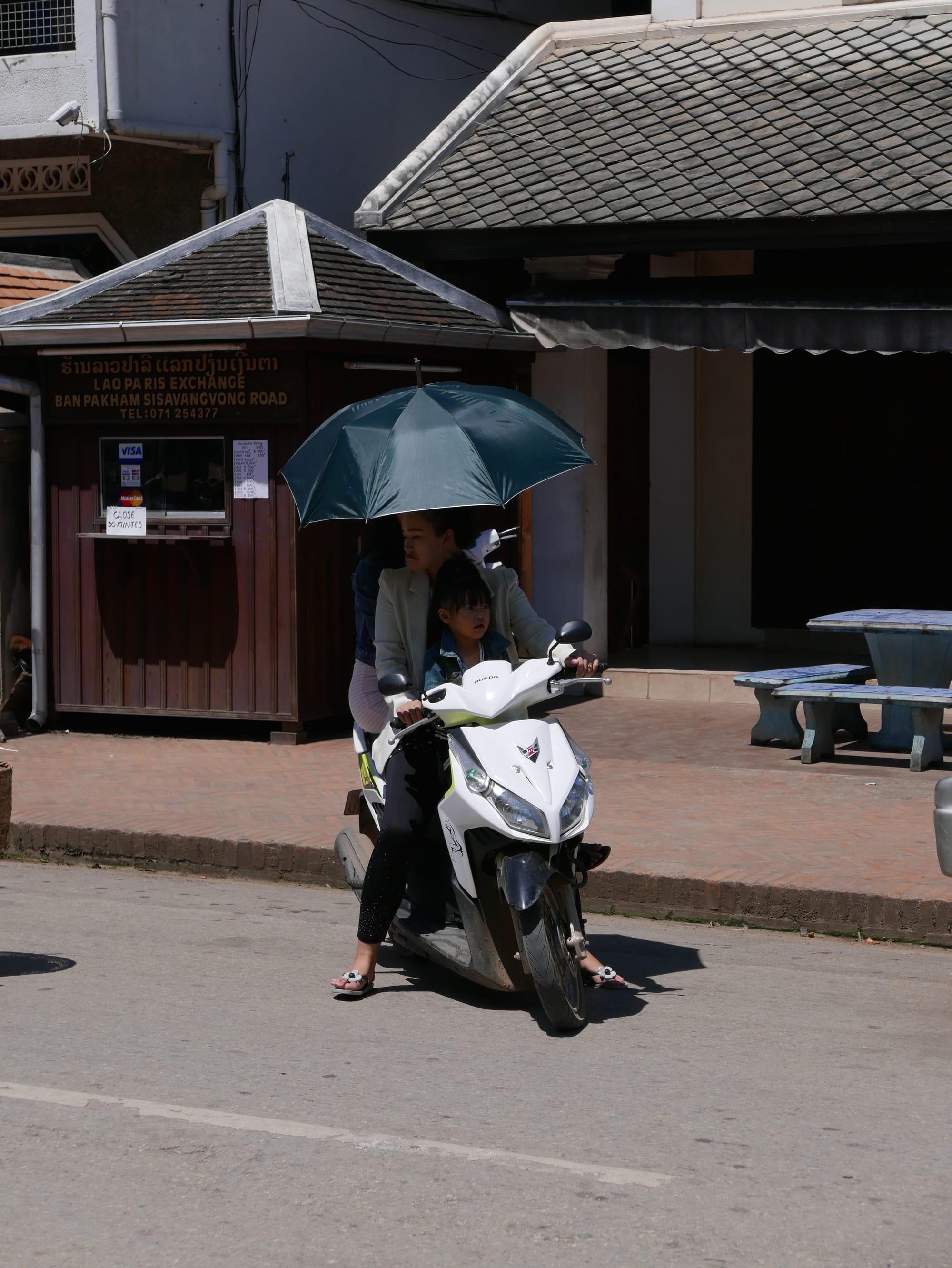 Photo by Author — motorbikes in Luang Prabang (ຫລວງພະບາງ/ຫຼວງພະບາງ), Laos — the complete set — passenger sitting sidesaddle holding an umbrella and the driver with a small child