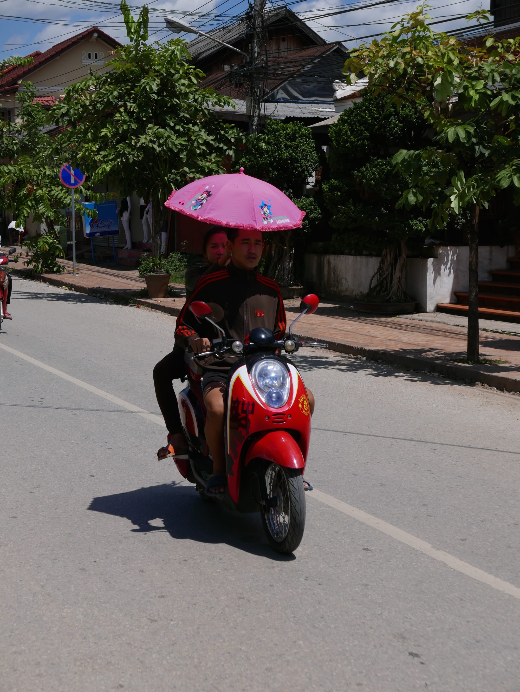 Photo by Author — motorbikes in Luang Prabang (ຫລວງພະບາງ/ຫຼວງພະບາງ), Laos — riding a motorbike with a passenger holding an umbrella